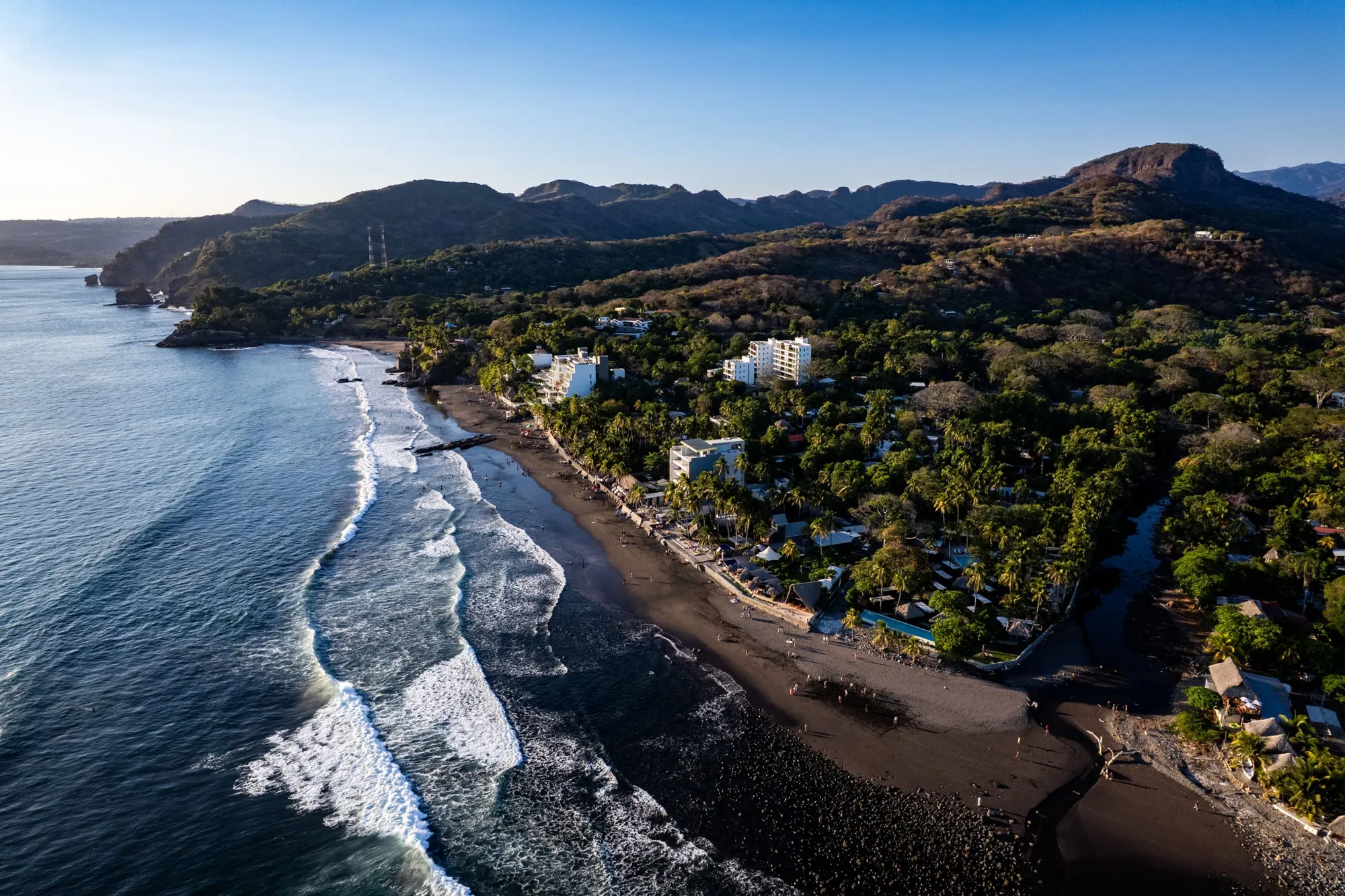Aerial view of El Zonte beach in La Libertad: a dark-sand bay, surf, and hotels and houses among trees below green hills.