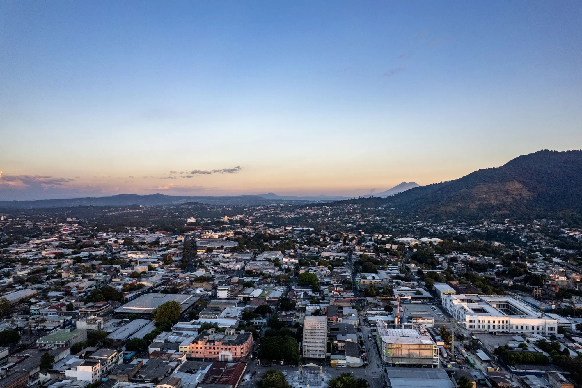 Aerial view of San Salvador at dusk, low-rise blocks stretching toward volcanoes on the horizon.